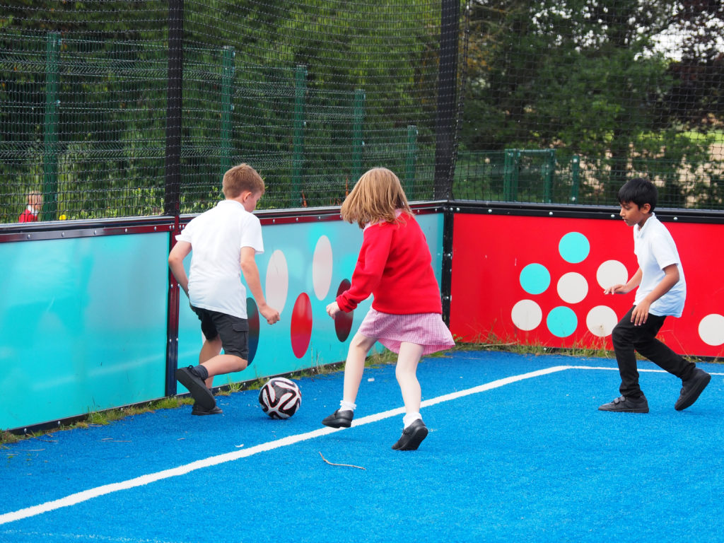 Two boys and a girl are shown playing Football together on an Astro Turf.