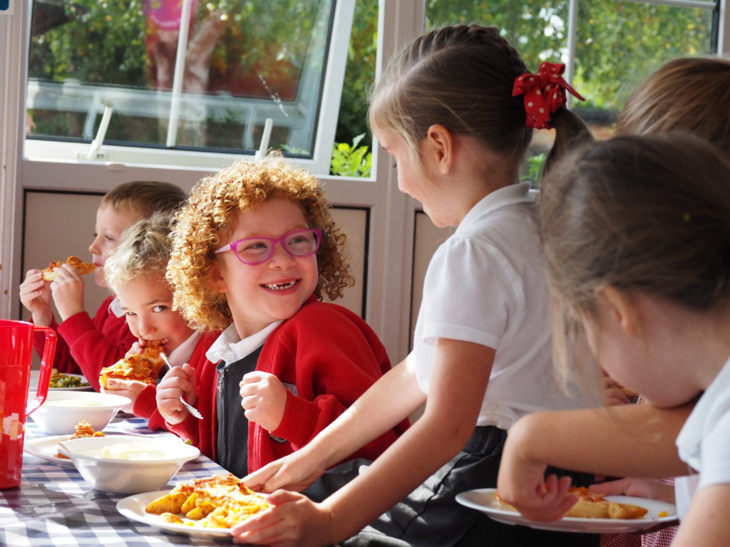 Six young pupils can be seen sitting together at a table, easting lunch and laughing with one another.
