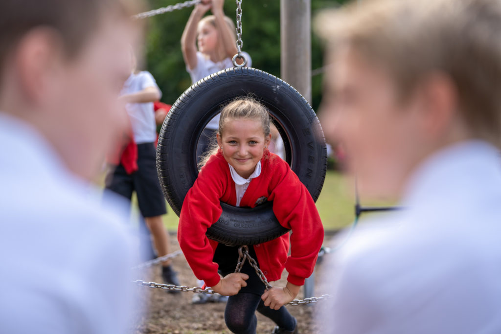 A young girl is seen hanging through a tyre attached to a climbing frame and smiling for the camera.