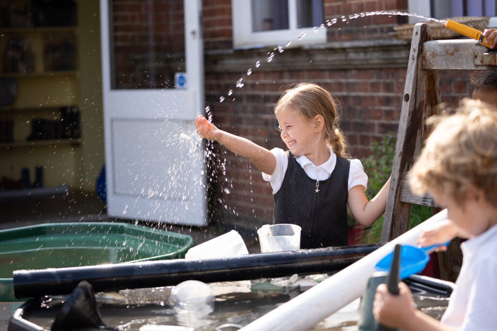 A female school student playing with water shooting from a hose pipe in an outdoor play area.
